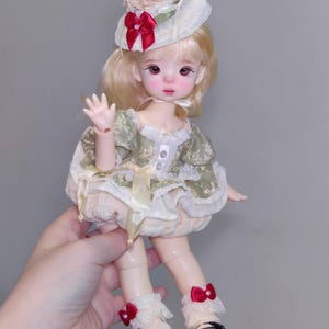 May include: A detailed doll with blonde hair, wearing a green and cream dress, a hat with a red bow and a cherry on top. The doll has black shoes with red bows and white lace socks. The doll is being held by a person.