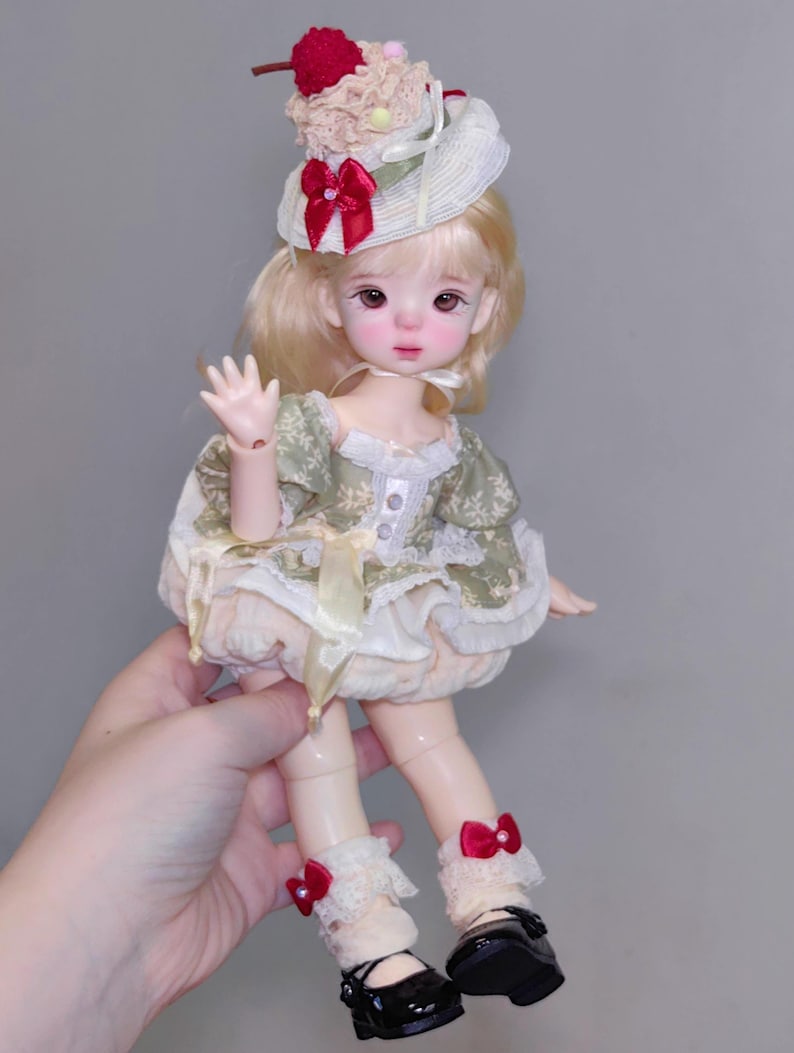 May include: A detailed doll with blonde hair, wearing a green and cream dress, a hat with a red bow and a cherry on top. The doll has black shoes with red bows and white lace socks. The doll is being held by a person.