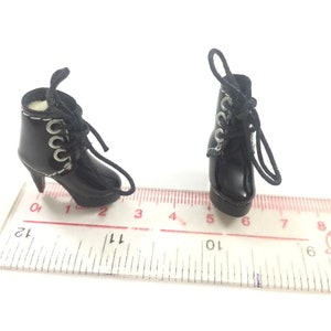 May include: A pair of black heeled boots with silver eyelets and black laces. The boots are on a white ruler with black markings.