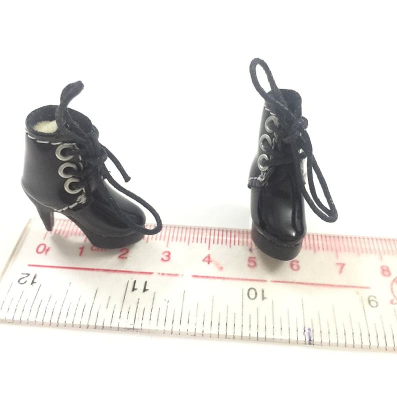 May include: A pair of black heeled boots with silver eyelets and black laces. The boots are on a white ruler with black markings.