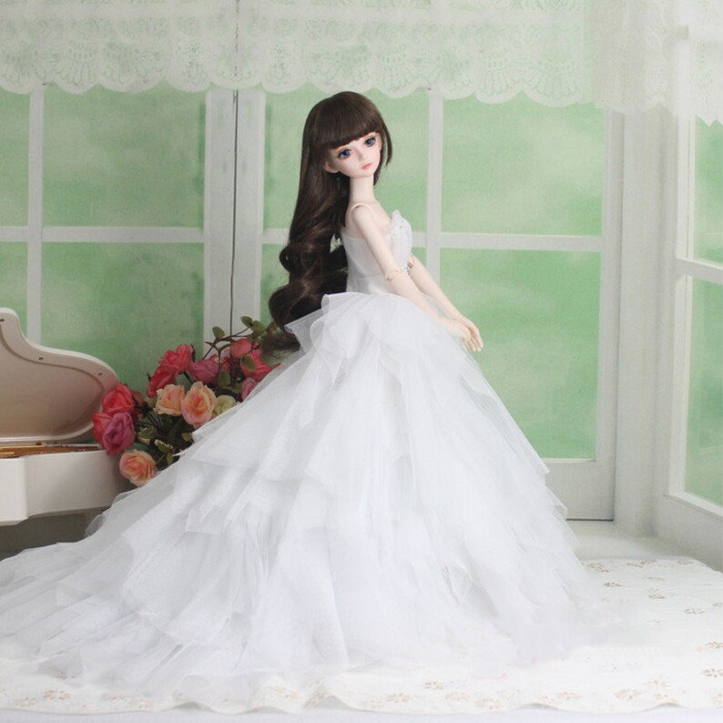 May include: A white bridal gown with a full skirt and layers of tulle. The doll is wearing a white dress with a sweetheart neckline and a fitted bodice. The doll has long brown hair and blue eyes.