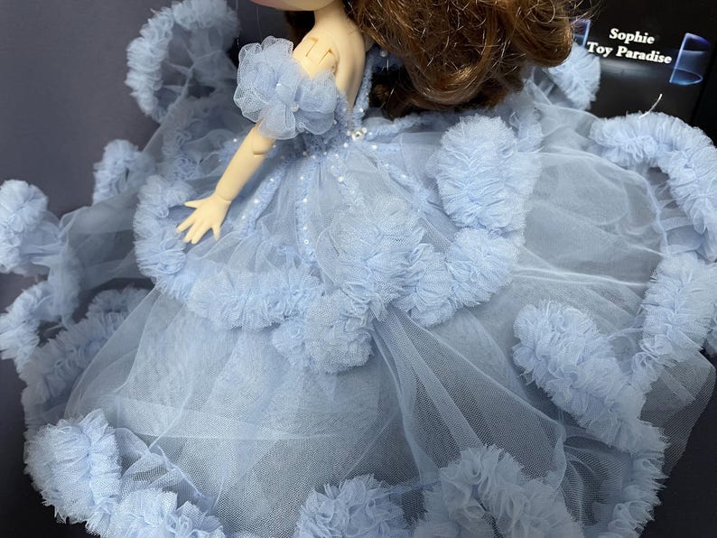 May include: A light blue tulle dress with ruffled trim and a beaded bodice. The dress is designed for a doll and has a long train.