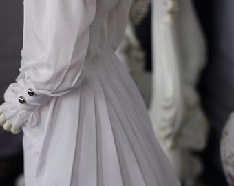May include: A white dress with a pleated skirt and long sleeves. The dress has a ruffled collar and silver buttons on the cuffs.