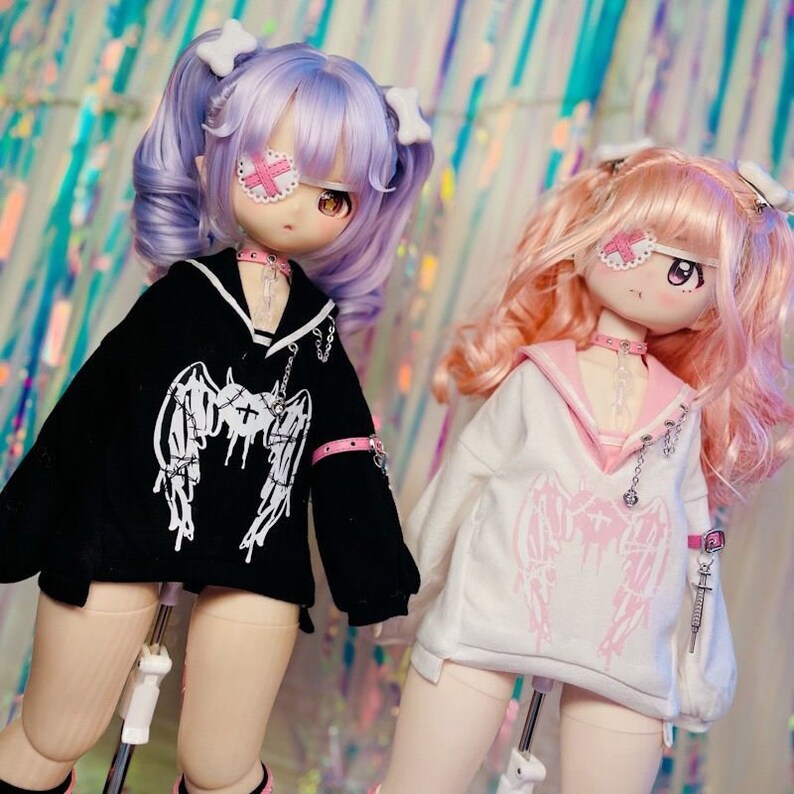 K&ouml;nnte beinhalten: Zwei Puppen mit langen, bunten Haaren. Eine Puppe tr&auml;gt ein schwarz-wei&szlig;es Outfit mit einem wei&szlig;en Augenpflaster. Die andere Puppe tr&auml;gt ein wei&szlig;-rosa Outfit mit einem rosa Augenpflaster. Beide Puppen haben Engelsfl&uuml;gel auf ihren Outfits.