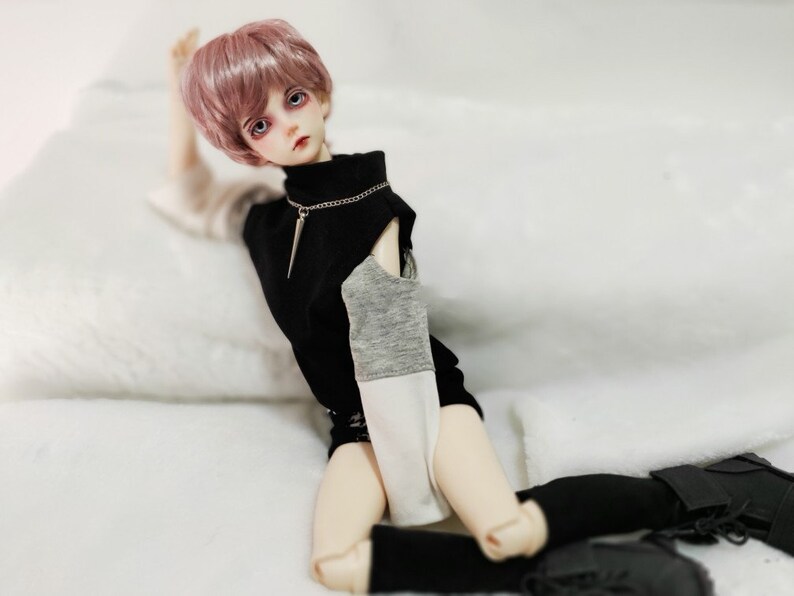 May include: A doll with pink hair wearing a black and grey shirt with a white undershirt, black pants, and black boots. The doll is sitting on a white surface.