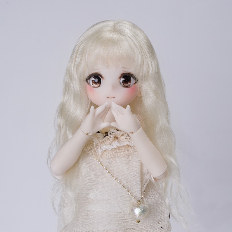 May include: A ball-jointed doll with long wavy platinum blonde hair, large brown eyes, and rosy cheeks. The doll wears a cream-colored sleeveless top with a delicate lace collar and a thin gold-toned chain necklace featuring a small heart pendant.
