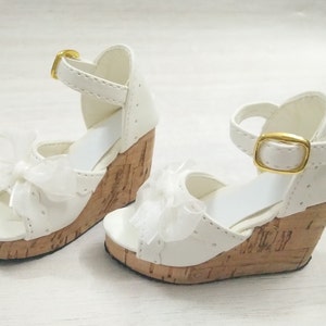 Peut inclure: Une paire de sandales compens&eacute;es blanches avec des talons en li&egrave;ge et une boucle dor&eacute;e. Les sandales ont un n&oelig;ud en ruban blanc &agrave; l'avant.