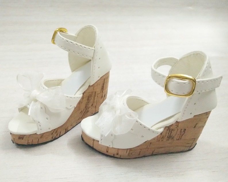 Peut inclure: Une paire de sandales compens&eacute;es blanches avec des talons en li&egrave;ge et une boucle dor&eacute;e. Les sandales ont un n&oelig;ud en ruban blanc &agrave; l'avant.