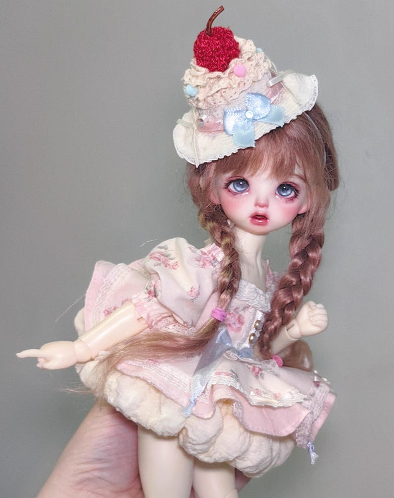 May include: A doll with blue eyes and long, braided brown hair, wearing a pink floral dress and a cupcake-shaped hat with a red cherry on top. The doll's dress has a layered skirt and lace trim.