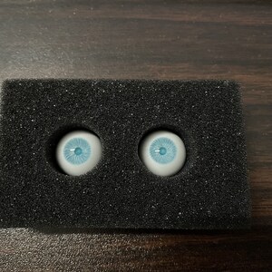 K&ouml;nnte beinhalten: Zwei blaue Glasaugen mit wei&szlig;en Iris und schwarzen Pupillen, in einer schwarzen Schaumstoffverpackung.