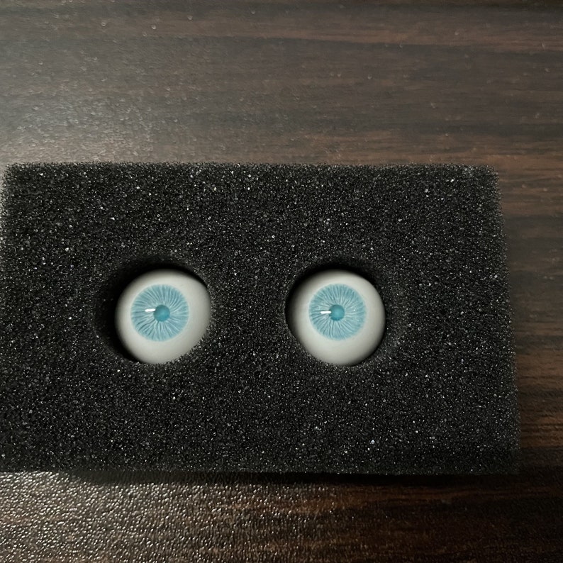 K&ouml;nnte beinhalten: Zwei blaue Glasaugen mit wei&szlig;en Iris und schwarzen Pupillen, in einer schwarzen Schaumstoffverpackung.