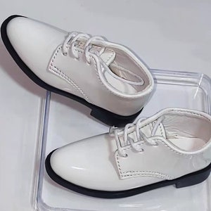 Op de afbeelding: Een paar witte lakleren veterschoenen met zwarte zolen.