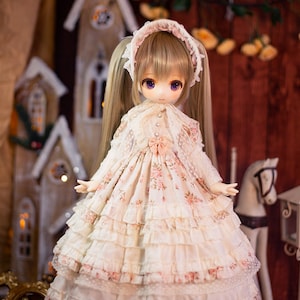 May include: A doll wearing a white dress with pink floral print and lace trim. The doll has long blonde hair in pigtails and purple eyes. The doll is standing in front of a wooden background with a miniature house and a white rocking horse.