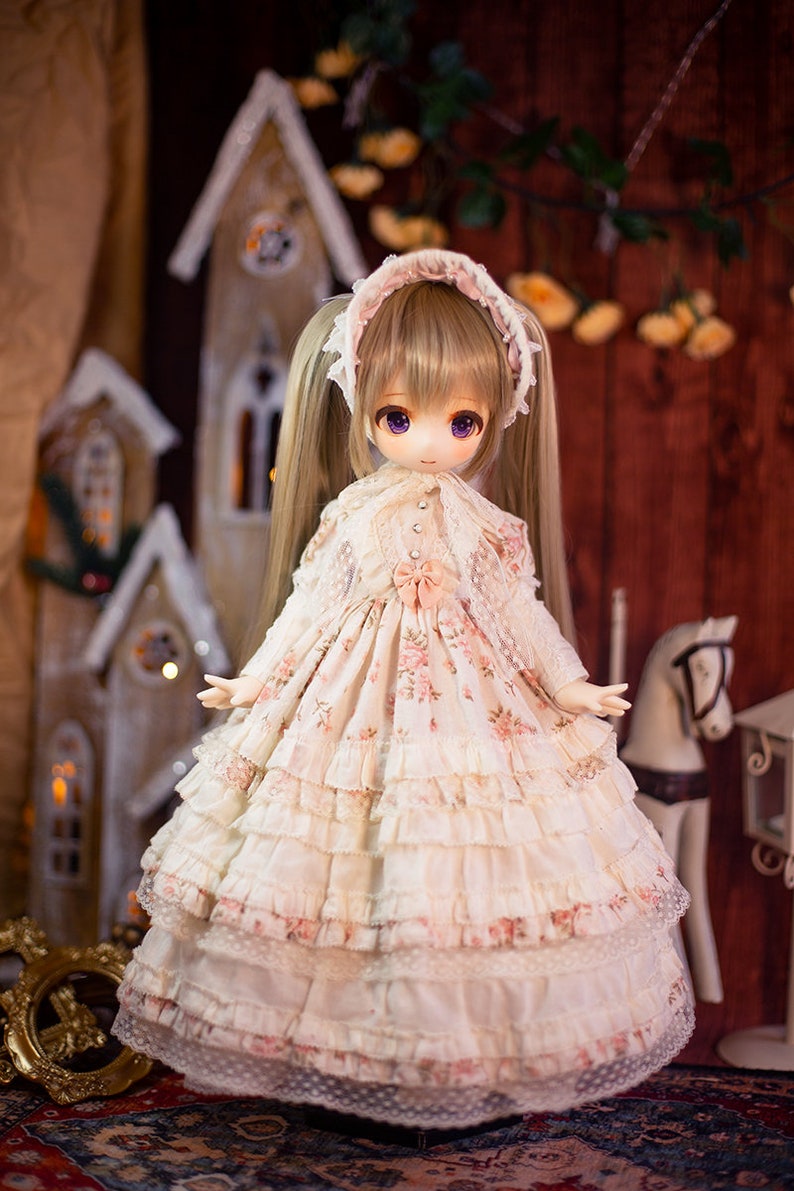 May include: A doll wearing a white dress with pink floral print and lace trim. The doll has long blonde hair in pigtails and purple eyes. The doll is standing in front of a wooden background with a miniature house and a white rocking horse.