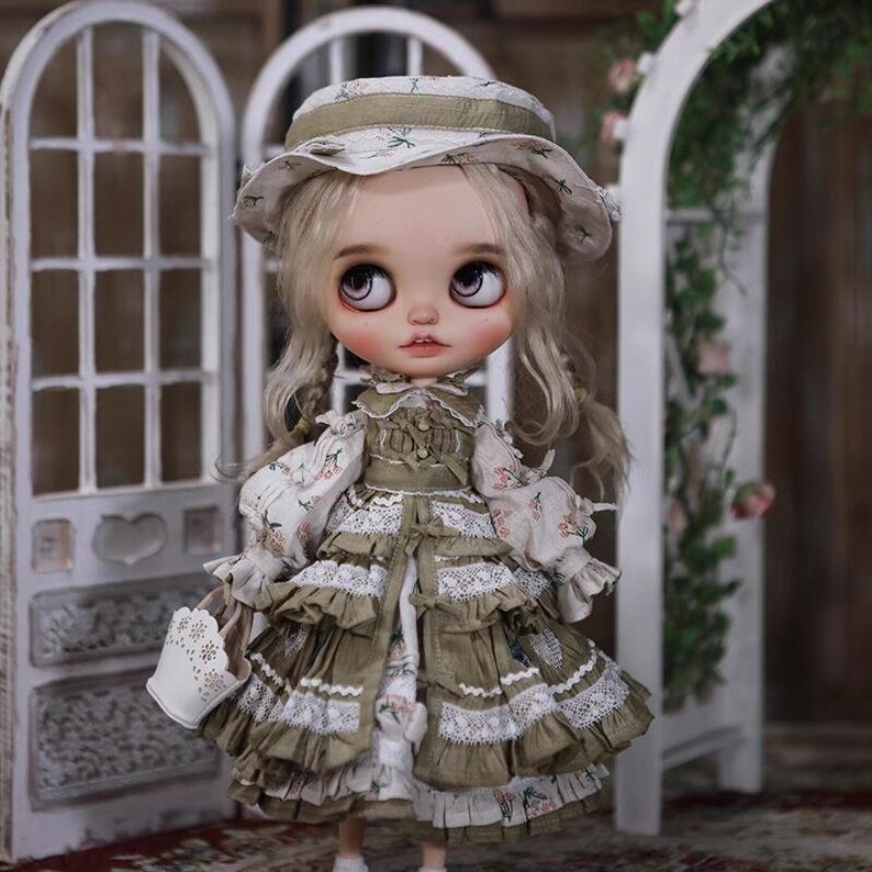 May include: A doll wearing a green and white dress with lace trim and a matching hat. The doll has blonde hair and blue eyes. The doll is holding a white basket.