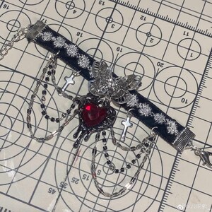 K&ouml;nnte beinhalten: Eine schwarze Choker-Kette mit silbernen Akzenten, mit einem tropfenden roten Herz-Anh&auml;nger, einem Schmetterlings-Anh&auml;nger und Kreuz-Anh&auml;ngern. Die Kette ist mit mehreren Ketten verziert.