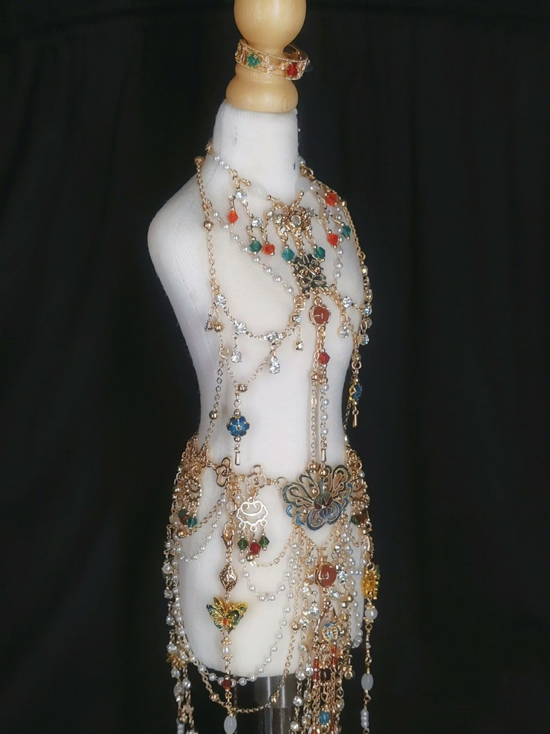 May include: A gold-toned body chain with multiple layers of chains, beads, and charms. The chain features a variety of colorful beads, including red, green, blue, and white. The charms include a butterfly, a flower, and a crescent moon.