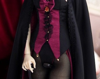 Op de afbeelding: Pop in een zwart en bordeauxrood kostuum met een cape. De outfit bevat een korset, strik en gouden accenten. De pop houdt een wandelstok vast en draagt ​​visnetkousen.