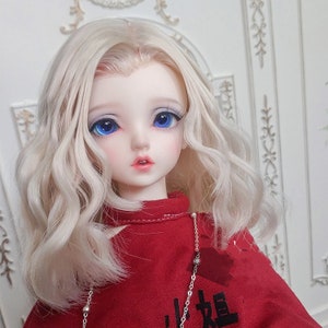 May include: A ball-jointed doll with wavy platinum blonde hair, large bright blue eyes, and pale skin. The doll wears a red long-sleeved top with a high collar and black Chinese characters printed on the front, accented by a thin silver chain necklace.
