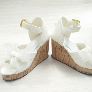 Peut inclure: Une paire de sandales compens&eacute;es blanches avec une plateforme en li&egrave;ge et un n&oelig;ud blanc &agrave; l'avant. Les sandales ont une boucle dor&eacute;e sur la bride de cheville.