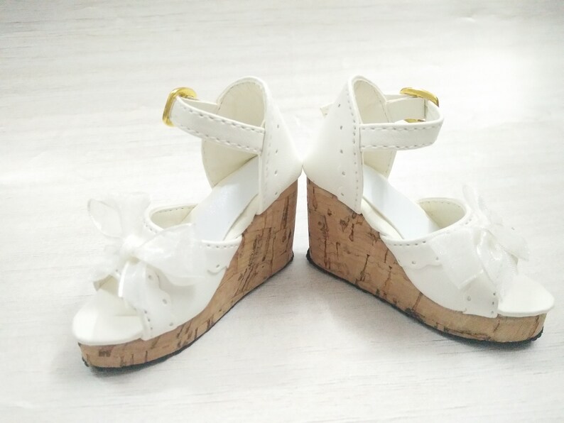 Peut inclure: Une paire de sandales compens&eacute;es blanches avec une plateforme en li&egrave;ge et un n&oelig;ud blanc &agrave; l'avant. Les sandales ont une boucle dor&eacute;e sur la bride de cheville.