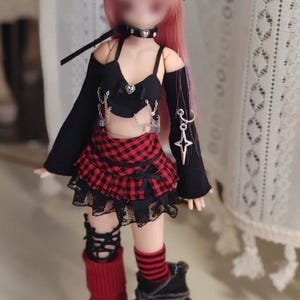 Op de afbeelding: Een pop in een zwarte outfit met een rode geruite rok, zwarte hoed en rood-zwarte beenwarmers. De pop heeft roze haar en draagt een zwarte choker. De outfit bevat een zwarte top met zilveren kettingen en een sterhanger.