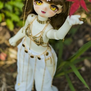 May include: A doll with long brown hair wearing a white outfit with gold accents and a gold chain headpiece. The doll is holding out its hand towards a red leaf.