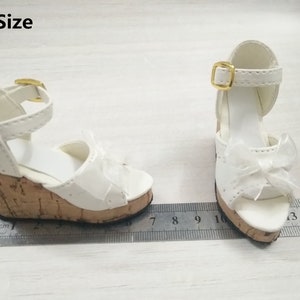 Peut inclure: Une paire de sandales compens&eacute;es blanches avec une plateforme en li&egrave;ge et une boucle dor&eacute;e. Les sandales ont un d&eacute;tail de n&oelig;ud sur le devant et sont de taille 1/3.