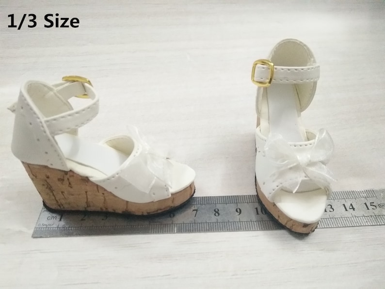 Peut inclure: Une paire de sandales compens&eacute;es blanches avec une plateforme en li&egrave;ge et une boucle dor&eacute;e. Les sandales ont un d&eacute;tail de n&oelig;ud sur le devant et sont de taille 1/3.