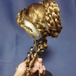 May include: A brown doll wig with a braided bun and a long, curly ponytail. The wig is displayed on a metal stand.
