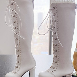 Pode incluir: Um par de botas brancas at&eacute; o joelho com design de cadar&ccedil;o e saltos pequenos. As botas t&ecirc;m uma borda ondulada na frente e um z&iacute;per na lateral. Uma guarni&ccedil;&atilde;o branca com babados adorna a parte superior das botas.