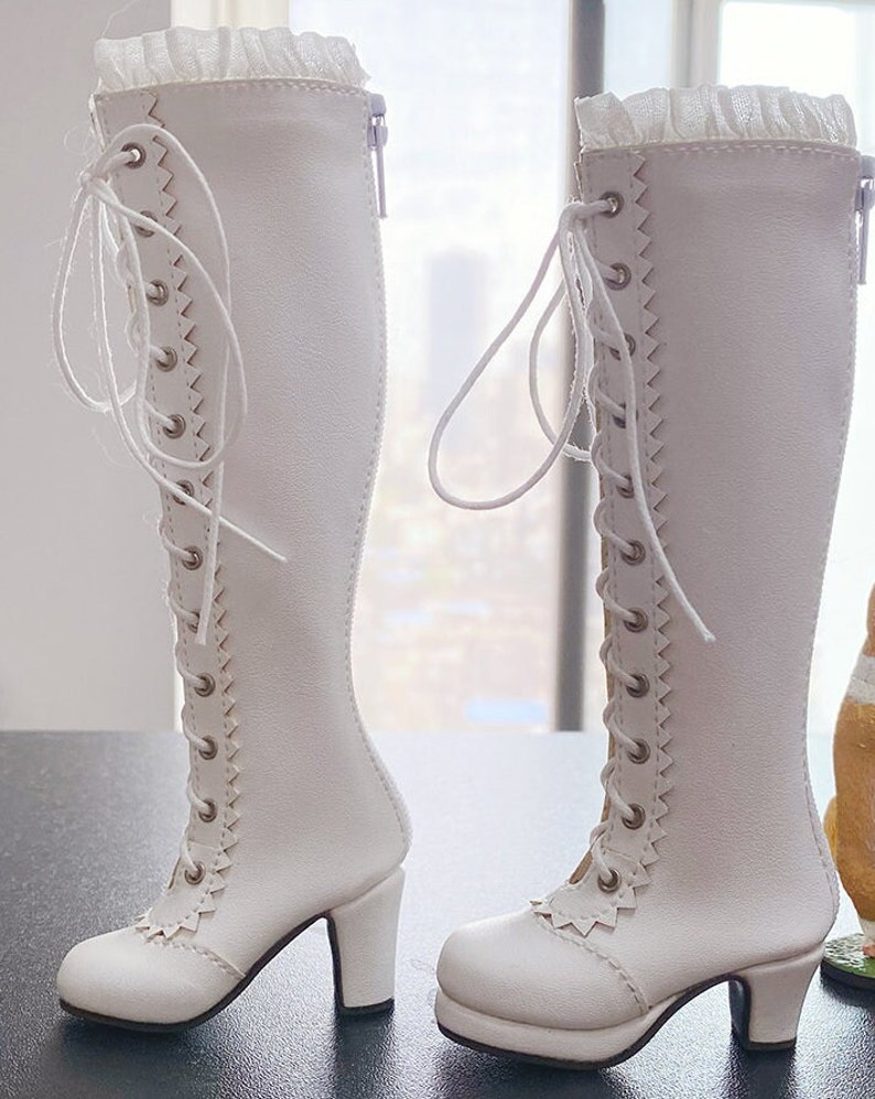 Pode incluir: Um par de botas brancas at&eacute; o joelho com design de cadar&ccedil;o e saltos pequenos. As botas t&ecirc;m uma borda ondulada na frente e um z&iacute;per na lateral. Uma guarni&ccedil;&atilde;o branca com babados adorna a parte superior das botas.