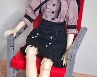 May include: A doll wearing a pink ruffled blouse with a black bow and a black skirt with white accents sits on a red chair with a gray frame.