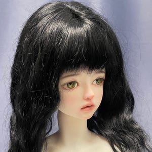 May include: A ball-jointed doll head featuring pale skin, light green eyes, and long, wavy black hair with bangs. The doll has a neutral facial expression with soft pink lips and defined eyelashes, capturing a detailed, realistic aesthetic.