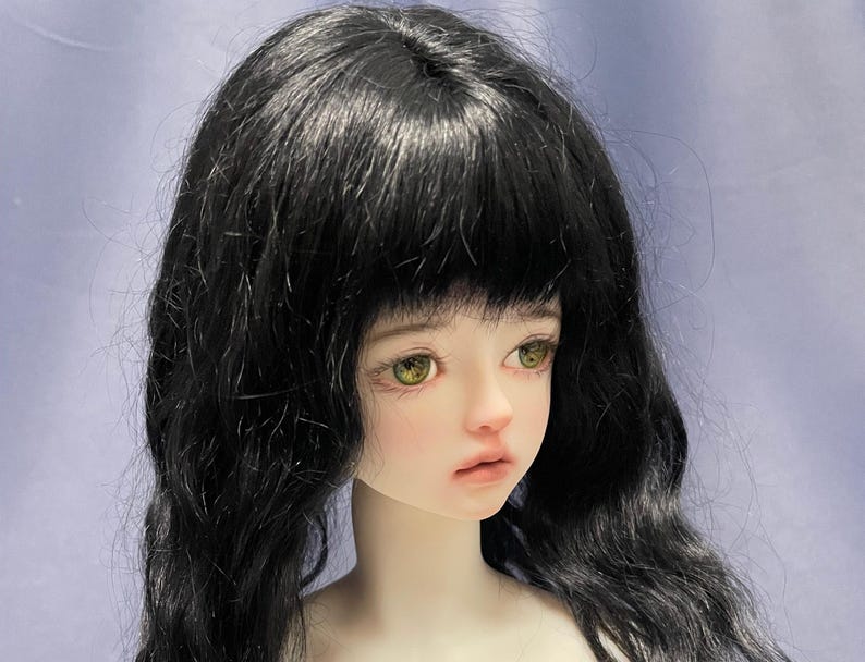 May include: A ball-jointed doll head featuring pale skin, light green eyes, and long, wavy black hair with bangs. The doll has a neutral facial expression with soft pink lips and defined eyelashes, capturing a detailed, realistic aesthetic.
