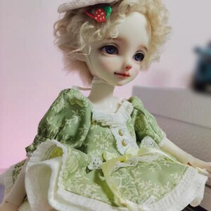 May include: A doll with a fair complexion, blue eyes, and blonde curly hair. The doll wears a green dress with a floral pattern, white lace trim, and a cream-colored skirt. A small hat with a strawberry and red bow sits atop the doll's head.