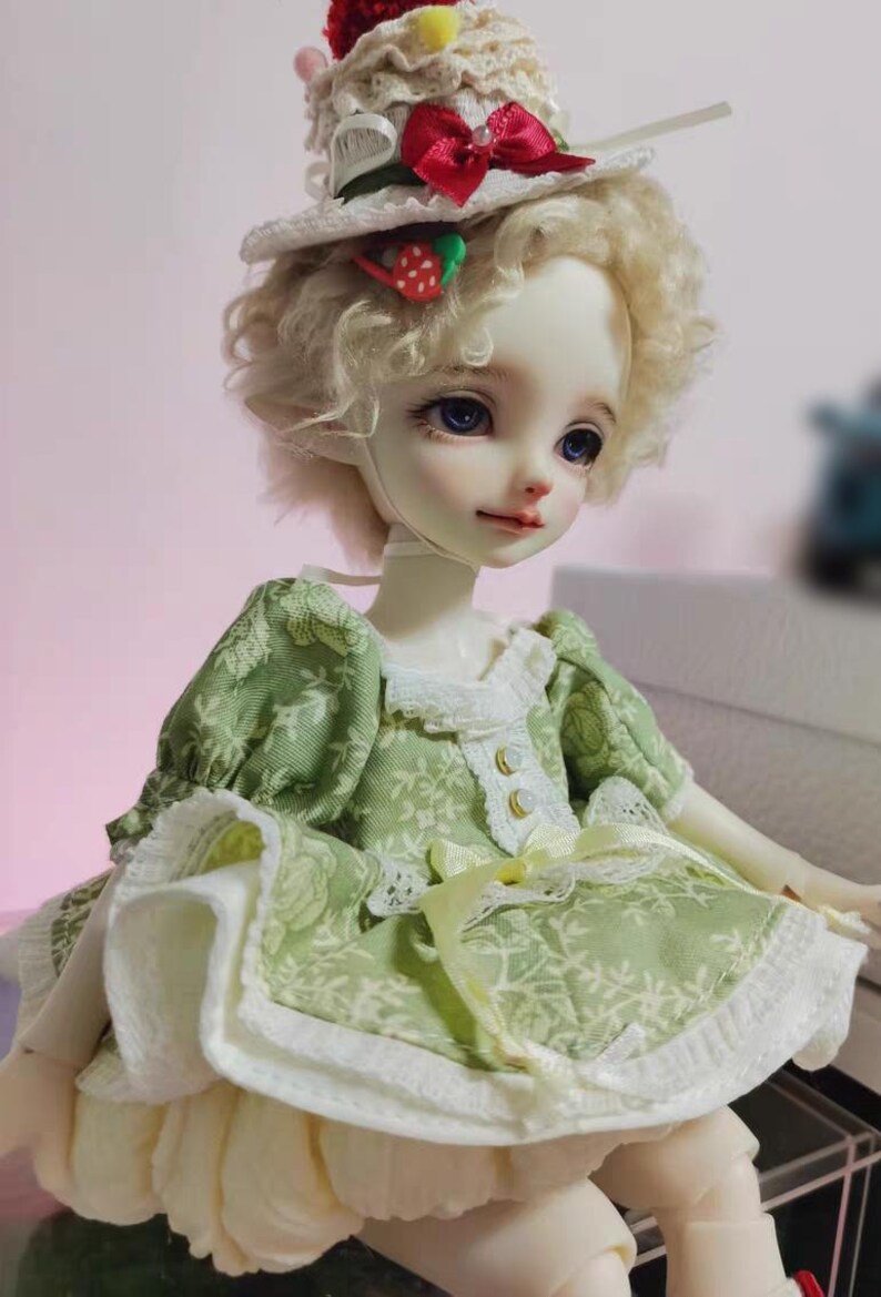 May include: A doll with a fair complexion, blue eyes, and blonde curly hair. The doll wears a green dress with a floral pattern, white lace trim, and a cream-colored skirt. A small hat with a strawberry and red bow sits atop the doll's head.