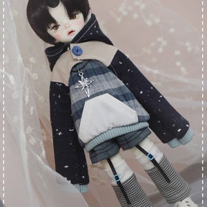 May include: A doll with black hair and a horn on its forehead is wearing a blue and white plaid hoodie with a white pocket and a blue button. The doll is also wearing black and white checkered pants and white shoes.