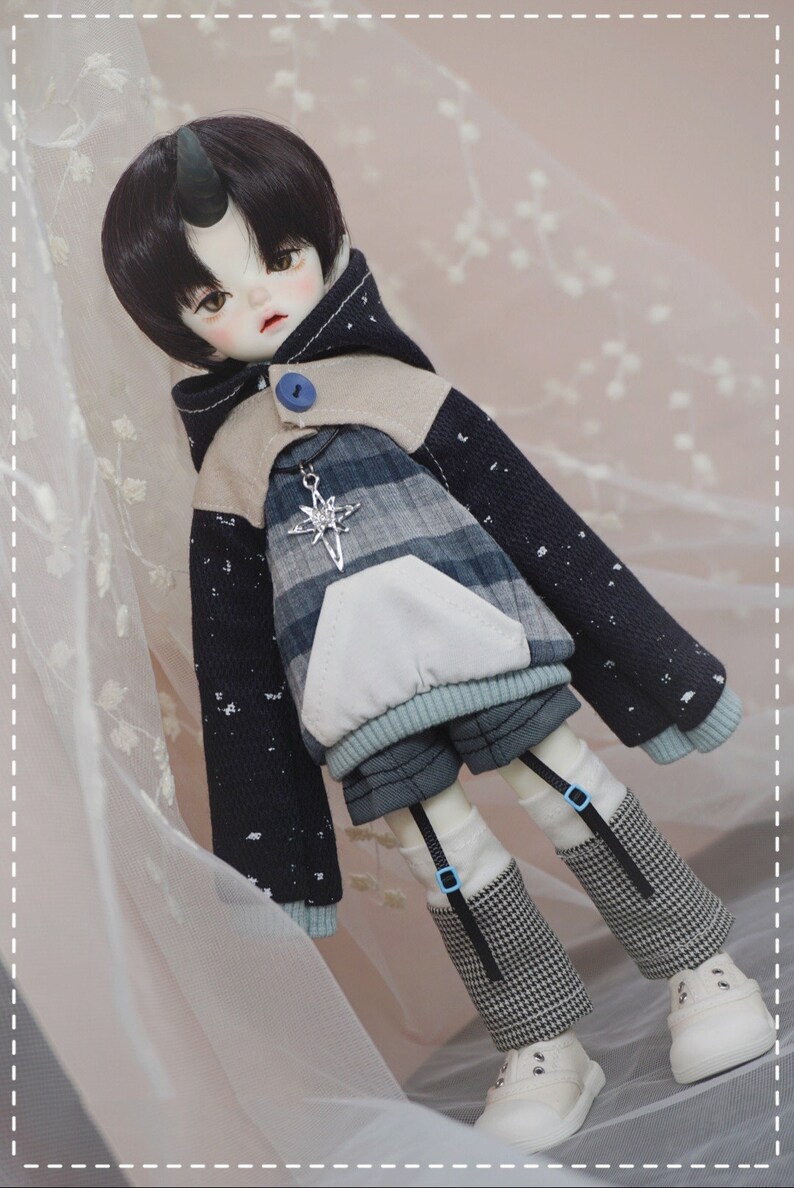 May include: A doll with black hair and a horn on its forehead is wearing a blue and white plaid hoodie with a white pocket and a blue button. The doll is also wearing black and white checkered pants and white shoes.