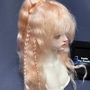 Puede incluir: Una muñeca con el pelo largo, ondulado y de color melocotón, peinado en una coleta con dos trenzas. El pelo se mantiene en su lugar con un lazo rosa claro. La muñeca tiene ojos verdes y lleva una blusa blanca.