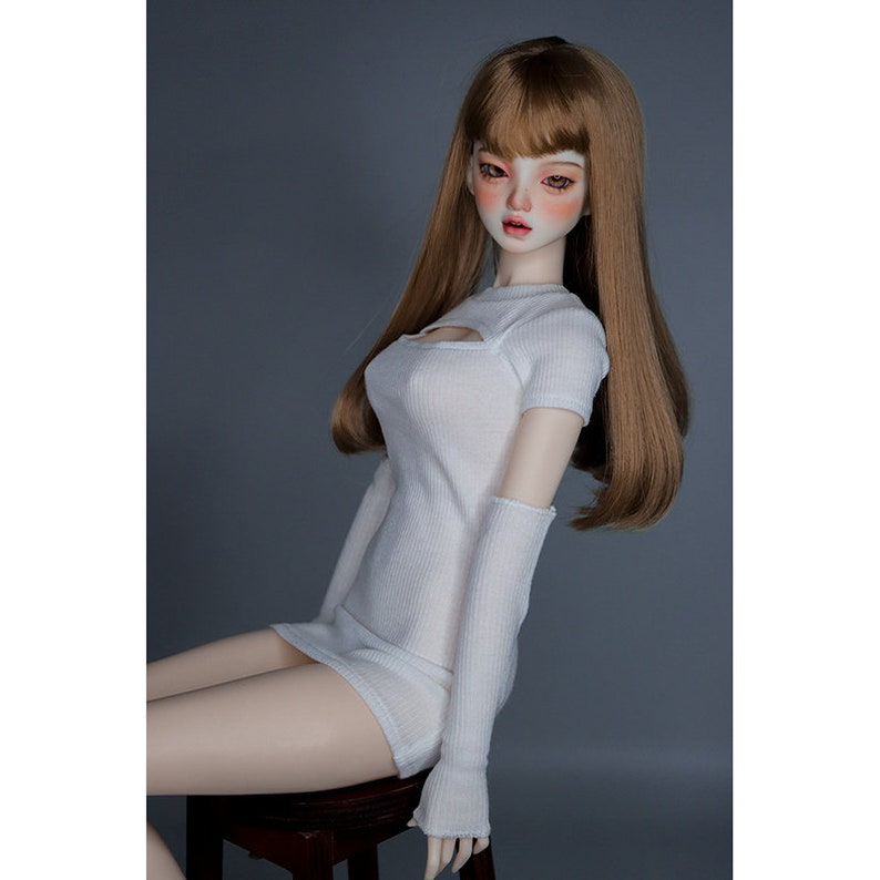 May include: A white doll wearing a white ribbed knit dress with a cutout detail at the neckline. The doll has long brown hair and is sitting on a wooden stool.