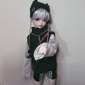 May include: A doll with white hair wearing a black and gray outfit with a black hat with cat ears. The doll is holding out its hand.