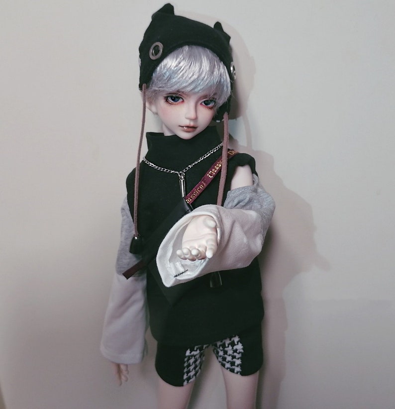 May include: A doll with white hair wearing a black and gray outfit with a black hat with cat ears. The doll is holding out its hand.