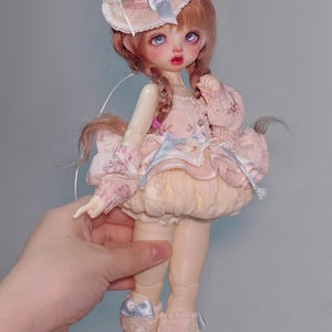 May include: A doll with a pink dress, a hat with a cherry on top, and light brown hair in braids. The doll is wearing a pink dress with lace and a light blue bow. The doll is wearing beige socks and white shoes.