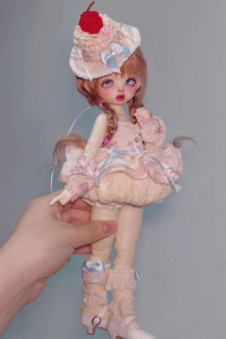 May include: A doll with a pink dress, a hat with a cherry on top, and light brown hair in braids. The doll is wearing a pink dress with lace and a light blue bow. The doll is wearing beige socks and white shoes.