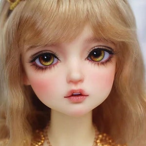 Peut inclure: Gros plan sur le visage d'une poup&eacute;e avec de longs cheveux blonds. La poup&eacute;e a de grands yeux jaunes brillants avec des pupilles brunes et un teint rose doux.