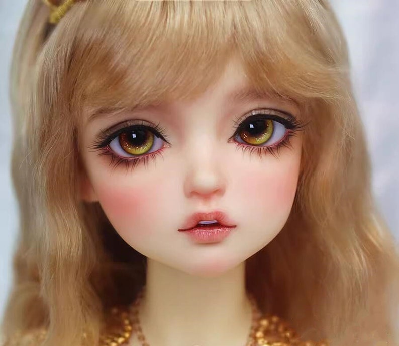 Peut inclure: Gros plan sur le visage d'une poup&eacute;e avec de longs cheveux blonds. La poup&eacute;e a de grands yeux jaunes brillants avec des pupilles brunes et un teint rose doux.