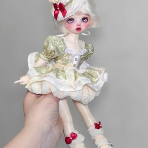 May include: A doll with pale skin, blue eyes, and white hair, wearing a green and cream-colored dress with a matching hat. The hat is adorned with a red bow and a decorative topping. The doll is wearing white shoes with a small heel and red bows.