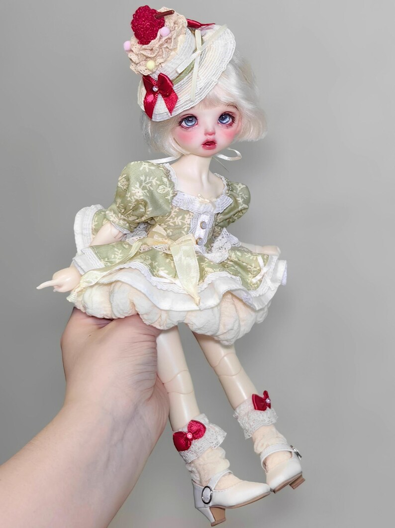 May include: A doll with pale skin, blue eyes, and white hair, wearing a green and cream-colored dress with a matching hat. The hat is adorned with a red bow and a decorative topping. The doll is wearing white shoes with a small heel and red bows.