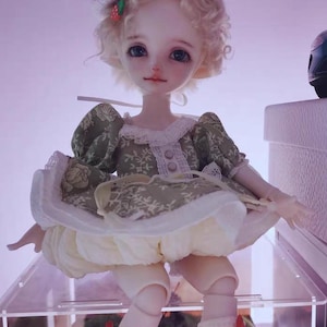 May include: A detailed doll with a whimsical design. The doll has a fair complexion, blonde curly hair, and is wearing a green dress with a white lace collar and a cream-colored skirt. A decorative hat with red accents tops the doll's head.
